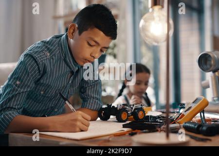 Zuerst Hausaufgaben, danach Spielzeit. Aufnahme eines hübschen Jungen, der zu Hause seine Hausaufgaben zur Robotik macht, mit seiner jüngeren Schwester im Hintergrund. Stockfoto