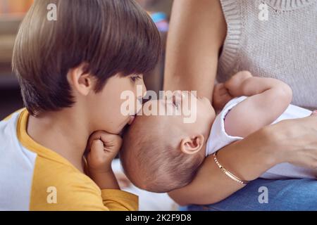 Der süßeste große Bruder, den es je gab. Aufnahme eines entzückenden kleinen Jungen, der seinen kleinen Bruder küsst, während seine Mutter ihn zu Hause kadelt. Stockfoto