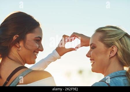 Wir reisen gerne, aber wir lieben uns mehr. Rückansicht von zwei schönen Freundinnen, die mit ihren Händen in der Natur eine Hitzeform bilden. Stockfoto