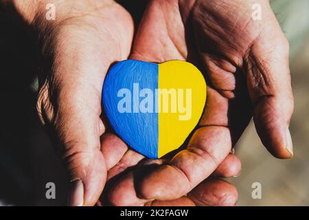 Seniorenhände oder Soldatenhände mit herzförmigem Stein bemalt mit ukrainischer Flagge Stockfoto