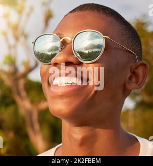 Nun, das nenne ich eine Aussicht. Aufnahme eines glücklichen jungen Mannes, der eine Sonnenbrille trägt, während er die Aussicht von draußen bewundert. Stockfoto