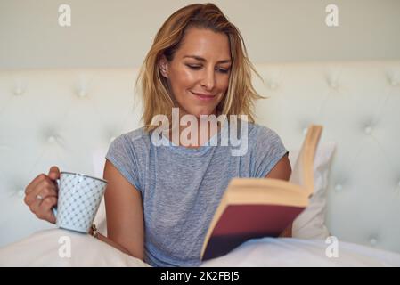 Dieses Buch hat mich Haken, Linie und Platine. Eine kurze Aufnahme einer attraktiven Frau, die zu Hause ein Buch liest und eine Tasse Kaffee im Bett trinkt. Stockfoto