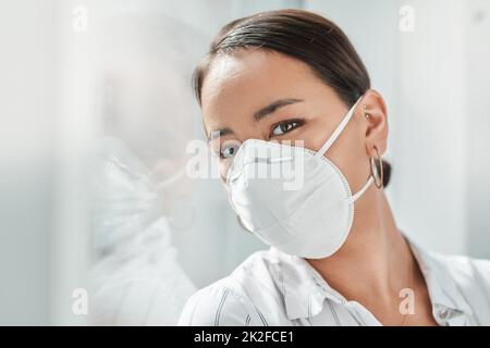 2020 das Jahr der Gesichtsmaske. Aufnahme einer maskierten jungen Geschäftsfrau, die sich in einem modernen Büro an einem Fenster lehnt. Stockfoto