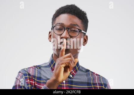 Sagen Sie niemandem ein Wort darüber. Porträt eines hübschen jungen Mannes, der mit seinem Finger auf den Lippen vor einem grauen Hintergrund posiert. Stockfoto