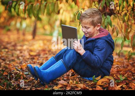 Modernes Kind. Ganzkörperaufnahme eines entzückenden kleinen Jungen mit einem Tablet, während er im Herbst im Freien sitzt. Stockfoto