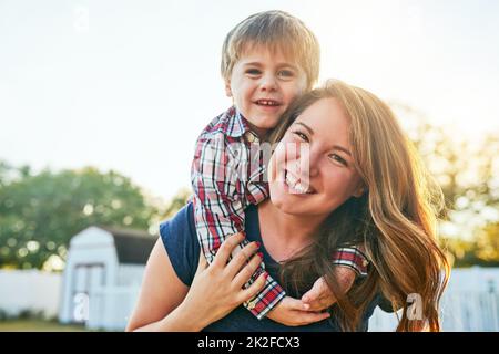 Es geht darum, diese lustigen Erinnerungen zu machen. Porträt einer Mutter, die ihrem entzückenden kleinen Sohn zu Hause eine Huckepack-Fahrt im Hinterhof gibt. Stockfoto