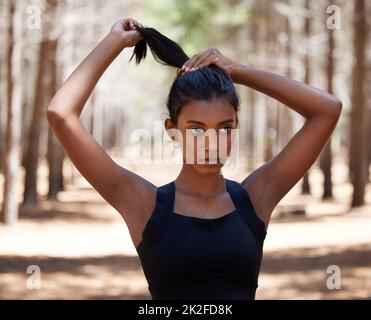 Haare auf, lasst uns trainieren. Aufnahme einer attraktiven jungen Frau, die allein draußen steht und ihre Haare vor ihrem Training bindet. Stockfoto