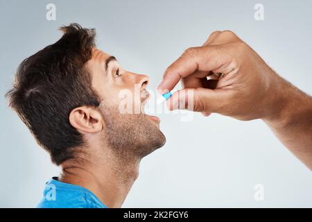 Weit geöffnet. Studioaufnahme eines sorglosen jungen Mannes, der von einer nicht erkennbaren Person eine Pille erhält, während er vor einem grauen Hintergrund steht. Stockfoto