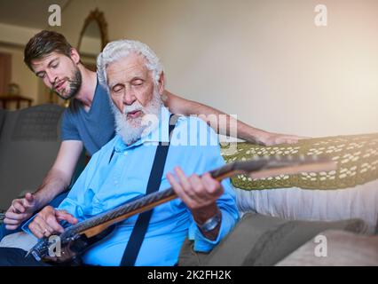 Du bist nie zu alt, um ein Rockstar zu werden. Aufnahme eines glücklichen jungen Mannes, der seinem älteren Großvater beibringt, zu Hause auf der Couch E-Gitarre zu spielen. Stockfoto