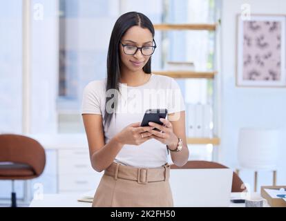 Kommunikation ist der Eckpfeiler ihres Geschäfts. Ausgeschnittene Aufnahme einer attraktiven jungen Frau, die eine SMS sendet, während sie in ihrem Büro steht. Stockfoto