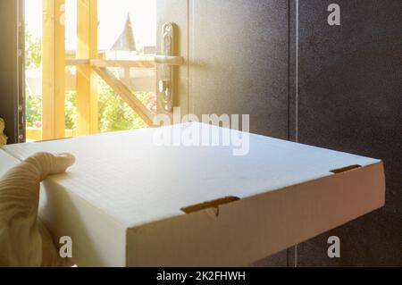 Nahaufnahme einer Pizzaschachtel aus Pappe, die Hand eines Kuriers in einem Gummihandschuh, mit einer Lebensmittellieferbox vor dem Hintergrund einer offenen Tür in einem Landhaus Stockfoto