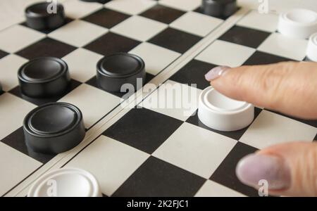 Die Hand einer Frau bewegt einen weißen Dame auf einem schwarz-weißen Spielfeld, das Konzept von Hobbys und Heimspielen Stockfoto