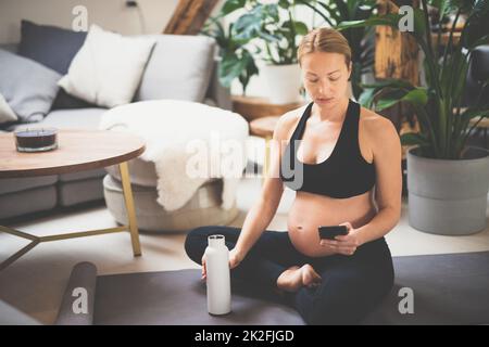 Junge glückliche und fröhliche schöne Schwangere, die eine Pause macht, feuchtigkeitsspendend, Trinkwasser aus der Flasche nach Hause Wohlbefinden Trainingsprogramm. Stockfoto