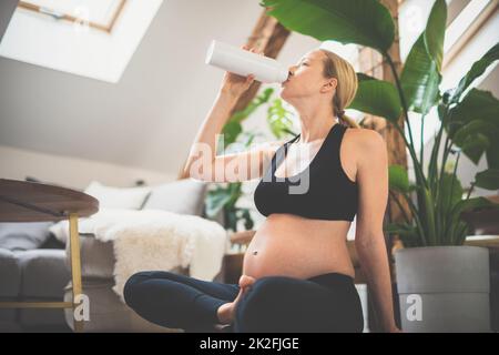 Junge glückliche und fröhliche schöne Schwangere, die eine Pause macht, feuchtigkeitsspendend, Trinkwasser aus der Flasche nach Hause Wohlbefinden Trainingsprogramm. Stockfoto