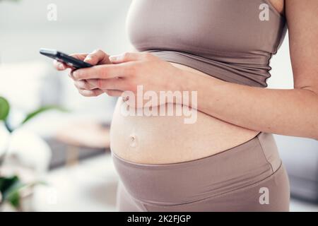 Nahaufnahme des schwangeren weiblichen Bauches. Frau, die eine Smartphone-App in der Wohnung hält und verwendet. Schwangerschaft, Technologie, Online-Shopping, Vorbereitung und Erwartungskonzept. Stockfoto