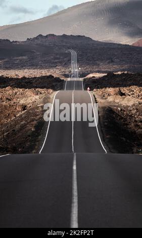 Endlose Straße auf einem Vulkan im Timanfaya-Nationalpark auf Lanzarote auf den Kanarischen Inseln mit einer durchgehenden Linie, schwarzen vulkanischen Felsen an der Seite und Vulkanen im Nebel im Hintergrund. Stockfoto