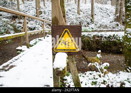 Rutschiges Warnschild im Wald, Holzbrücke mit Schnee bedeckt, Wintersaison, Vorsicht vor Gefahr, im Freien Stockfoto