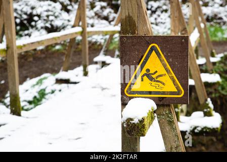 Rutschiges Warnschild im Wald, Holzbrücke mit Schnee bedeckt, Wintersaison, Vorsicht vor Gefahr, im Freien Stockfoto