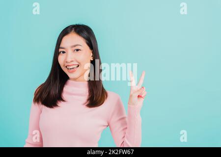 Junge asiatische, schöne lächelnde Frau tragen kiefergerecht geformte Silikon-Zahnhalter auf den Zähnen, die V-Zeichen zeigen, Siegesfinger isoliert auf blauem Hintergrund, nach abnehmbaren Zahnspangen. Zahnhygienisches Konzept Stockfoto