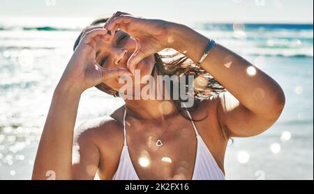 Ich liebe Menschen, die den Sommer lieben. Porträt einer schönen jungen Frau, die mit ihren Händen am Strand eine herzförmige Geste macht. Stockfoto