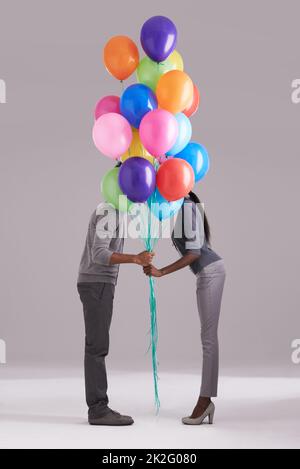 Einen Kuss hinter den Ballons stehlen. Studioaufnahme eines Paares, das sich hinter einem Haufen Ballons versteckt. Stockfoto