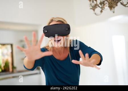 Verschwimmen der Grenzen zwischen Fantasie und Realität. Eine kurze Aufnahme einer reifen Frau, die zu Hause ein VR-Headset trägt. Stockfoto