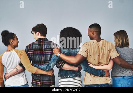 Unsere Freunde werden zu unserer Familie. Rückansicht eines Studios mit einer vielfältigen Gruppe junger Menschen, die sich vor grauem Hintergrund umarmen. Stockfoto