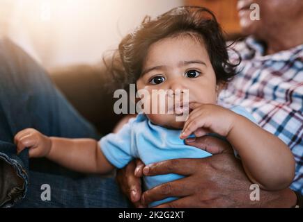 Ich versuche herauszufinden, was los ist. Porträt eines fröhlichen kleinen Jungen, der tagsüber auf dem Schoß seines Vaters sitzt und in die Kamera schaut. Stockfoto