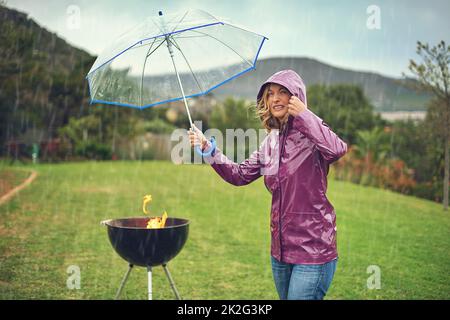 Nur das mutige Grillen im Regen. Aufnahme einer Frau, die einen Regenschirm hält, während sie versucht, im Regen zu grillen. Stockfoto