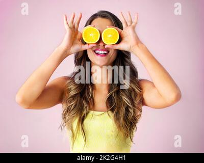 Schaut auf die helle Seite. Studioaufnahme einer jungen Frau, die zwei Hälften einer Orange vor ihren Augen hält. Stockfoto