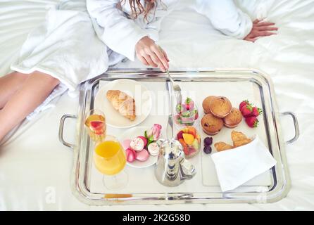 Beginnen Sie Ihren Morgen richtig. Die Aufnahme einer unkenntlichen Frau, die auf ihrem Hotelbett ein gesundes Frühstück genießt, ist aus einem hohen Winkel zu sehen. Stockfoto