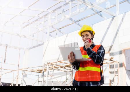 Eine Mitarbeiterin einer asiatischen Ingenieurin, die auf einer Baustelle arbeitet, verwendet einen Laptop und spricht mit dem Radio Stockfoto