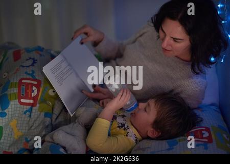 Er hat immer seine Flasche und ein Buch vor dem Schlafengehen. Ein kurzer Schuss eines kleinen Jungen, der im Bett liegt, während seine Mutter eine Schlafenszeit-Geschichte liest. Stockfoto