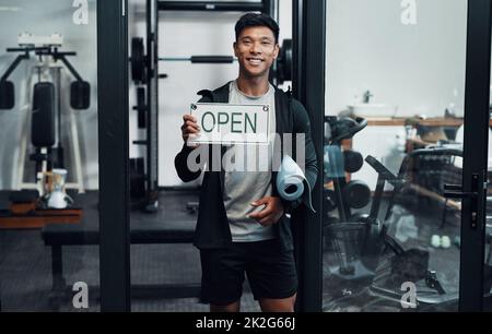 Sie sind jederzeit zwischen 9 und 5 Uhr willkommen. Verkürztes Porträt eines gutaussehenden jungen Fitnesstrainers, der ein Schild hochhält, auf dem "OFFEN" steht, während er in einem Fitnessstudio steht. Stockfoto