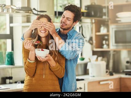 Raten Sie mal, wer. Aufnahme eines anhänglichen jungen Mannes, der die Augen seiner Freundinnen bedeckt, während sie in ihrer Küche frühstückt. Stockfoto