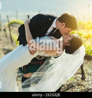 Was für ein atemberaubender Kuss. Aufnahme einer fröhlichen Braut und eines Bräutigams, die sich tagsüber draußen neben den Weinbergen einen Kuss teilen. Stockfoto