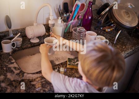 Hausarbeiten sind eine lebenswichtige Lebenskompetenz für Kinder zu lernen. High-Angle-Aufnahme eines kleinen Jungen, der in der Küche zu Hause Geschirr wascht. Stockfoto