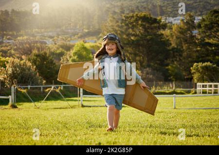 Fliegen auf den Flügeln der Phantasie. Aufnahme eines niedlichen kleinen Jungen, der einen Pilotenhut und eine Brille trägt, während er draußen mit Pappflügeln spielt. Stockfoto