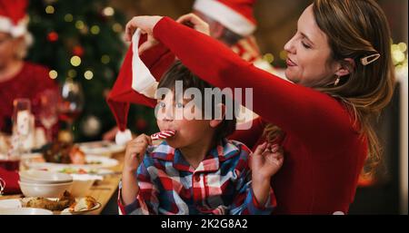 Der Weihnachtsmann hat Ihnen etwas geschickt. Aufnahme einer jungen Frau, die ihren Sohn während einer Weihnachts-Dinner-Party zu Hause mit dem Hut anlegt. Stockfoto