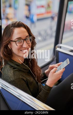 Ich kann SMS schreiben, wenn ich nicht fahren muss. Hochwinkelporträt einer attraktiven jungen Frau, die Musik hört, während sie in einem Bus sitzt. Stockfoto