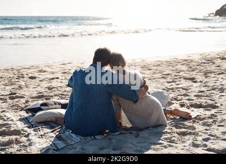 Es gibt nichts, was ein Sonnenuntergang am Strand nicht beheben kann. Rückansicht eines Paares mittleren Alters, das am Strand sitzt. Stockfoto