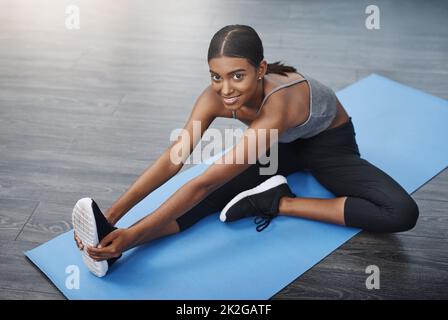 Mit sich selbst zufrieden zu sein, ist unübertroffen. Aufnahme einer wunderschönen jungen Frau, die zu Hause beim Sitzen und Stretching auf ihrer Sportmatte lächelt. Stockfoto