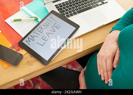 Jetzt ist es nicht mehr lange her Eine Schwangerin, die mit einer Tablette sitzt und die Worte Mutterschaftsurlaub auf dem Bildschirm hat. Stockfoto