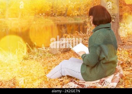 Frau, die auf Karli sitzt und in der Nähe des Teiches im wunderschönen Herbstpark ein Buch liest Stockfoto