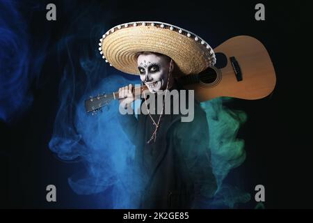 Junger Mann mit bemaltem Schädel auf dem Gesicht für Mexikos Day of the Dead (El Dia de Muertos) und Gitarre vor dunklem Hintergrund Stockfoto