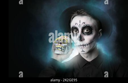 Junger Mann mit gemaltem Gesicht und Zuckerschädel für Mexikos Tag der Toten (El Dia de Muertos) vor dunklem Hintergrund Stockfoto