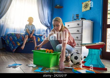 Die Spielzeit ist vorbei. Aufnahme einer Mutter, die Spielzeug wegpackt, während ihr Sohn im Hintergrund sitzt. Stockfoto