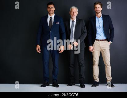 Drei ist die magische Zahl für den Erfolg. Studioportrait von drei Geschäftsleuten, die vor schwarzem Hintergrund stehen. Stockfoto