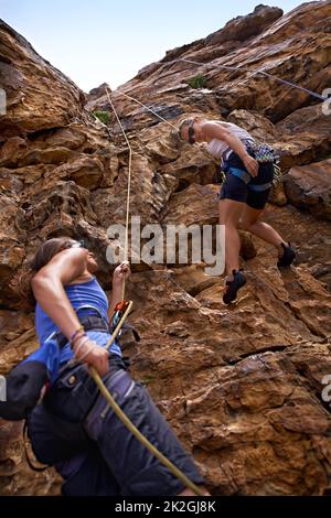 Es geht um Vertrauen - Sichermachen. Zwei weibliche Bergsteiger, die eine Felswand erklimmen. Stockfoto