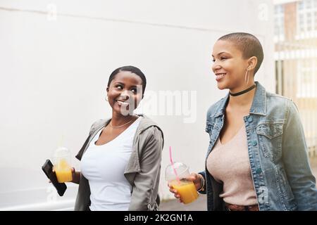 Zusammen durch dick und dünn. Aufnahme von zwei jungen Frauen, die in der Stadt lachend spazieren gehen, während sie ihre kühlen Getränke halten. Stockfoto